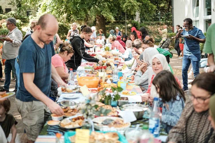 Menschen sitzen an einem Tisch im Freien. Auf dem Tisch stehen sehr viele unterschiedliche Gerichte aus verschiedenen Regionen der Welt.