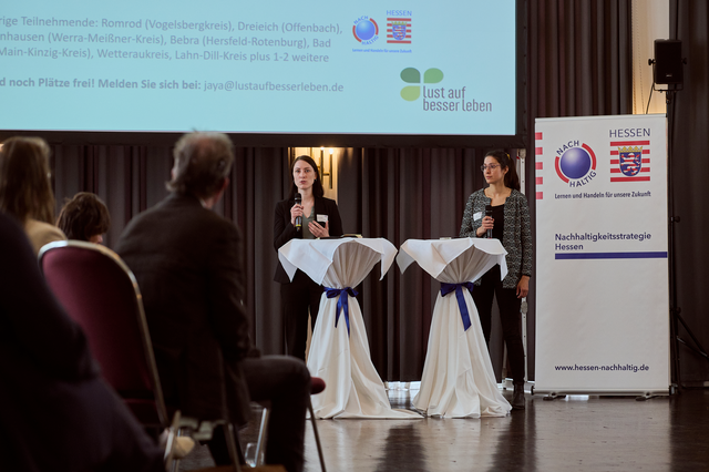 Zwei Frauen sprechen an Podiumstischen vor einem Publikum