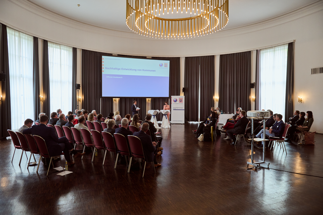 Foto in einen Raum hinein, in dem ein Publikum auf Stühlen sitzt, eine Präsentation gezeigt wird und zwei Personen vorne stehen und in Mikrofone sprechen